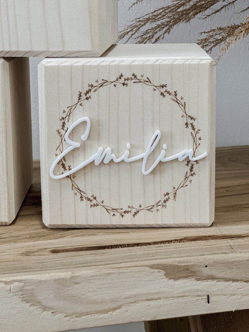 May include: A wooden block with the name "Emilia" in white script. The name is centered within a wreath design. The block is light-colored wood, and the wreath is a darker brown color. The block is sitting on a wooden surface.