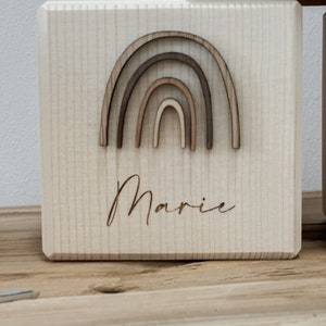 May include: A wooden block with a carved rainbow design in shades of brown. The name "Marie" is scripted below the rainbow. The block is made of light-colored wood and has a smooth finish. The block is sitting on a wooden surface.