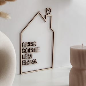 May include: Wooden house-shaped wall art with the names CHRIS, SOPHIE, LEV, and EMMA. The house outline is light brown with a dark brown border and a heart on the chimney. A beige candle and vase are in the background.