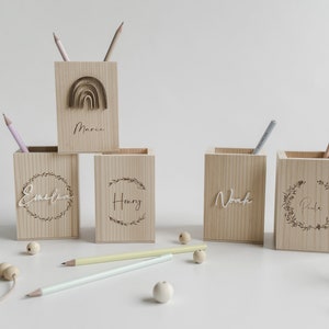 Puede incluir: Cinco porta lápices de madera con nombres personalizados. Los porta lápices están hechos de madera natural y tienen un color marrón claro. Los nombres están grabados en la parte delantera de cada porta lápices. Los nombres son: Emmeline, Henry, Noah y Riley. El porta lápices del centro tiene un diseño de arcoíris.