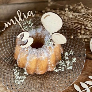 Puede incluir: Un adorno de pastel de madera con el nombre "Emilia" y el número "3" en él. El adorno se coloca en un pastel con glaseado blanco y un plato de madera. También hay decoraciones de madera en forma de globos y hojas.