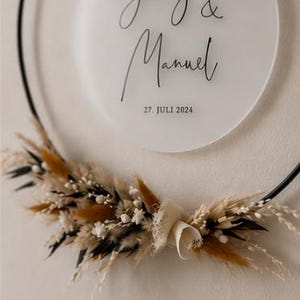 May include: A circular black metal hoop featuring a frosted acrylic disc with the black printed text Jenny & Manuel and the date 27. JULI 2024. The lower portion of the hoop is decorated with a cluster of dried grasses, white flowers, and brown foliage.