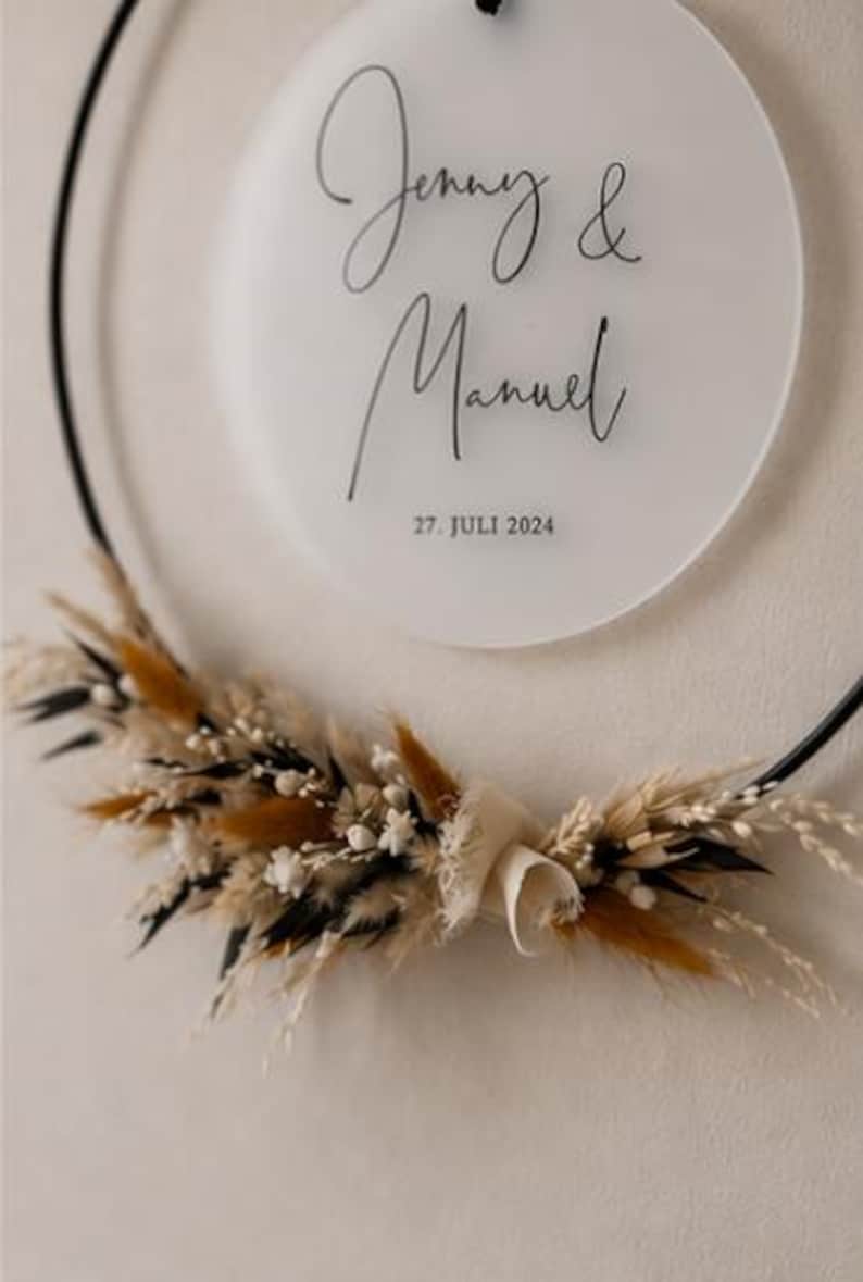 May include: A circular black metal hoop featuring a frosted acrylic disc with the black printed text Jenny & Manuel and the date 27. JULI 2024. The lower portion of the hoop is decorated with a cluster of dried grasses, white flowers, and brown foliage.