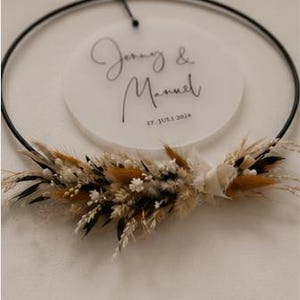 May include: A circular black metal hoop featuring a suspended frosted acrylic disc with the text Jenny & Manuel and 27. JULI 2024. The bottom of the hoop is decorated with a cluster of dried grasses, small white flowers, and dark foliage.