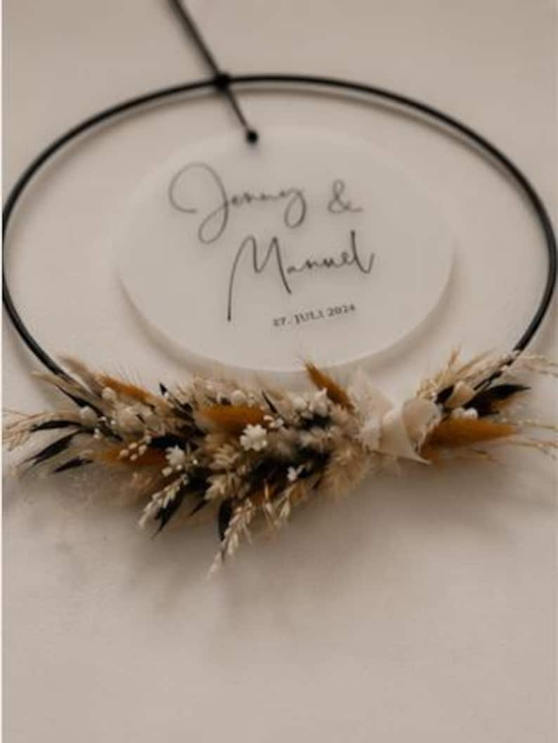 May include: A circular black metal hoop featuring a suspended frosted acrylic disc with the text Jenny & Manuel and 27. JULI 2024. The bottom of the hoop is decorated with a cluster of dried grasses, small white flowers, and dark foliage.