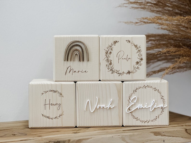 May include: Wooden blocks with names and decorative elements. One block features a rainbow, while others display names within floral wreaths. The names "Marie", "Paula", "Henry", "Noah", and "Emilia" are visible.