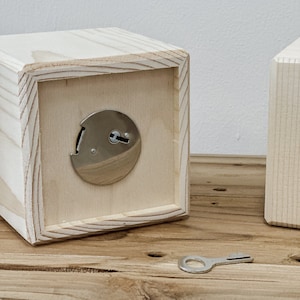 May include: A light-colored wooden box with a silver lock and key. The box is cube-shaped with a recessed front panel. The key is lying on a wooden surface. The box is made of unfinished wood.