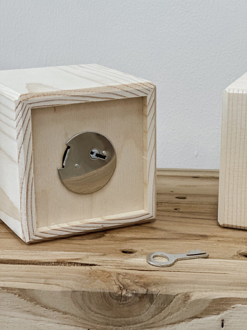 May include: A light-colored wooden box with a silver lock and key. The box is cube-shaped with a recessed front panel. The key is lying on a wooden surface. The box is made of unfinished wood.