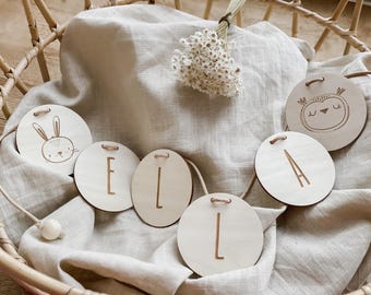 Peut inclure: Guirlande en bois avec des disques ronds en bois. Certains disques ont des lettres, d'autres ont une illustration de lapin et de hibou. La guirlande est présentée sur un tissu en lin beige à l'intérieur d'un panier en osier.