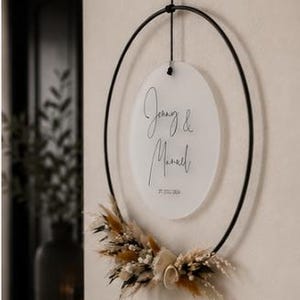 May include: A circular black metal hoop hanging from a black cord, featuring a frosted acrylic disc suspended in the center. The disc displays the names Jenny & Manuel and the date 27.07.2024. Dried beige and brown grasses decorate the bottom.