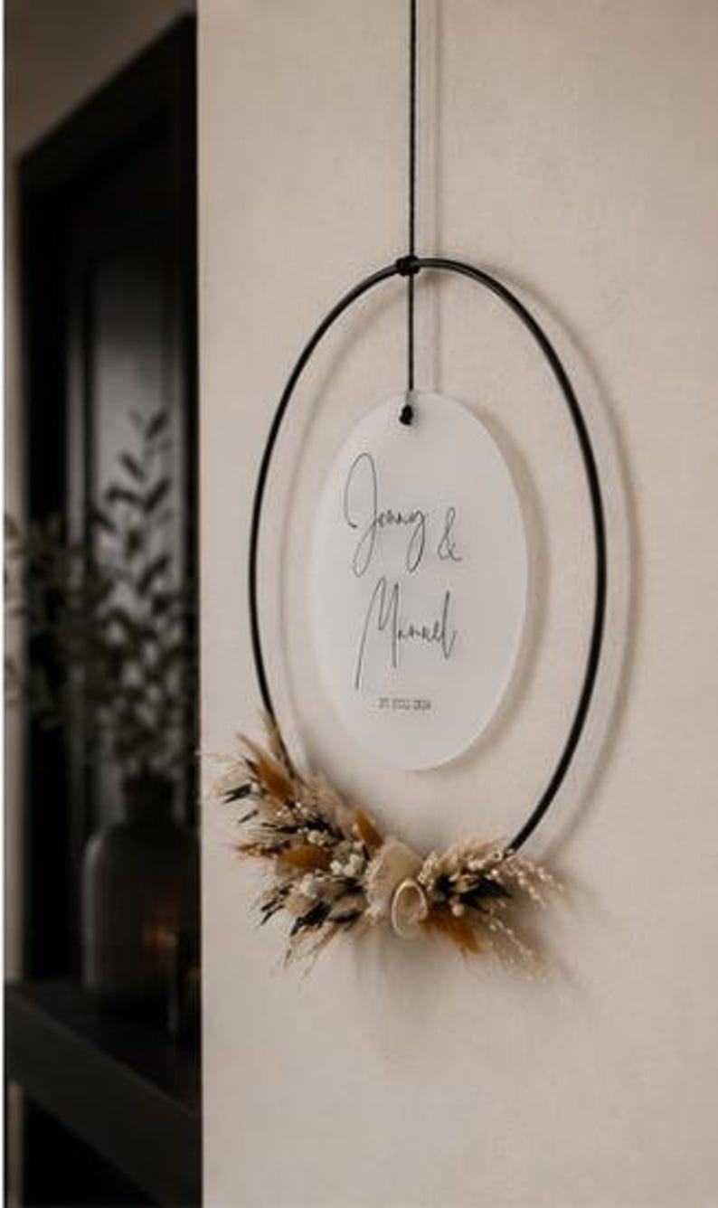 May include: A circular black metal hoop hanging from a black cord, featuring a frosted acrylic disc suspended in the center. The disc displays the names Jenny & Manuel and the date 27.07.2024. Dried beige and brown grasses decorate the bottom.