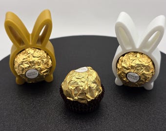 May include: An Easter display with two bunny-shaped holders, one gold and one white, each holding a Ferrero Rocher chocolate. A third chocolate sits between them. The chocolates are wrapped in gold foil.