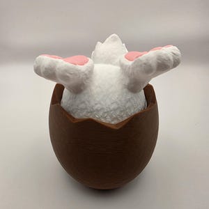 May include: A white bunny with pink paw pads emerges from a brown eggshell. The bunny's fluffy body is visible, with its paws extended. The eggshell has a textured appearance, creating a whimsical Easter-themed decoration.