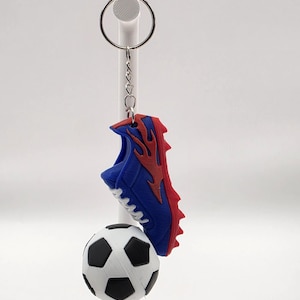 May include: A keyring featuring a blue and red football boot charm with a flame design and white laces, attached to a silver metal ring. Below the boot hangs a small, spherical football charm with a classic black and white pentagon pattern.