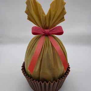 May include: A golden-coloured, wrapped gift, tied with a red bow and ribbon. The gift sits in a brown, fluted cup. The wrapping is gathered at the top, creating a decorative effect. The overall presentation is festive and elegant.