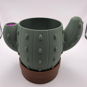May include: A sage green cactus-shaped planter with a textured surface and raised bumps. The planter features a brown base and a small, bright pink flower detail on one of the two side arms. The main body is cylindrical with a wide, open top.