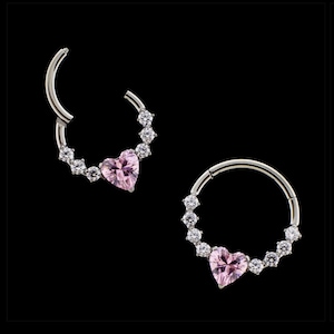Op de afbeelding: Twee zilverkleurige neusringen met een roze hartvormige edelsteen in het midden. Elke ring is omgeven door kleine, heldere edelstenen. De ringen staan tegen een zwarte achtergrond.
