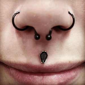 May include: Close-up of a person's face with multiple piercings. The person has a septum piercing with a black barbell, a nostril piercing with a black hoop, and a labret piercing with a black coffin-shaped stud.