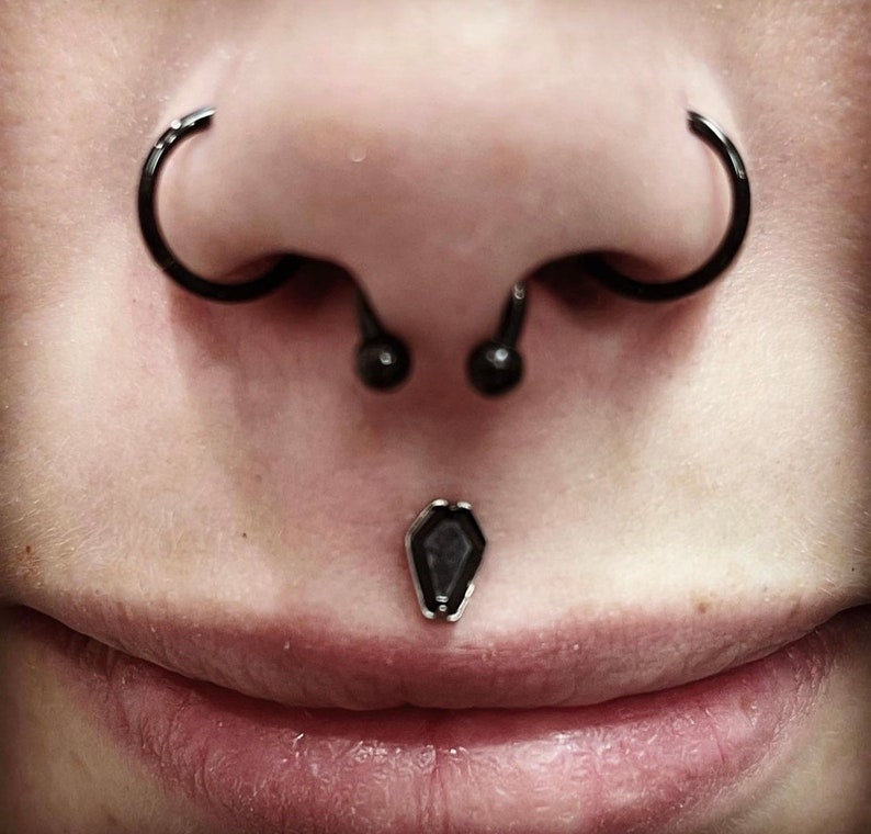 May include: Close-up of a person's face with multiple piercings. The person has a septum piercing with a black barbell, a nostril piercing with a black hoop, and a labret piercing with a black coffin-shaped stud.