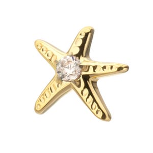 May include: A gold-toned starfish charm featuring a polished metallic finish and a single clear, round faceted gemstone set in the center. The five arms of the starfish are decorated with a series of small, circular indentations along the surface.