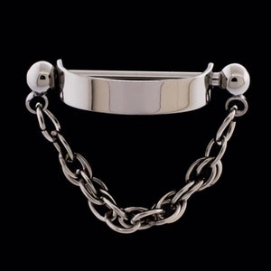 May include: A silver-colored body jewelry piece with a chain. The jewelry features a curved bar with a flat center section and rounded ends. A chain hangs from each end of the bar. The background is black.