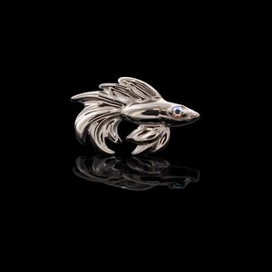 Può includere: Una spilla in metallo color argento lucido a forma di pesce combattente, con pinne fluenti e una piccola pietra blu incastonata come occhio. L'oggetto presenta una superficie liscia e riflettente su uno sfondo nero uniforme.