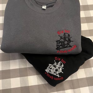 Op de afbeelding: Twee sweatshirts met geborduurde ontwerpen. Het bovenste sweatshirt is donkergrijs met een zwart-wit geborduurd ontwerp van een ridder met zwaarden en de tekst "Tis but a flesh wound". Het onderste sweatshirt is zwart met een wit geborduurd ontwerp van een ridder met zwaarden en de tekst "Tis but a flesh wound".