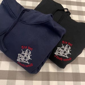 Puede incluir: Dos sudaderas con capucha, una azul marino y otra negra, con un diseño bordado en blanco de un caballero con espadas y el texto "Tis but a flesh wound".