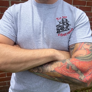 Puede incluir: Una camiseta gris con cuello redondo con un gráfico de un caballero y el texto "Tis but a flesh wound" en rojo. La camiseta está hecha de un material suave.