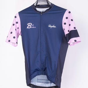 Può includere: Maglia da ciclismo blu navy con maniche a pois rosa. La maglia ha una cerniera a tutta lunghezza e le parole "Bonk Brothers" e "Rapha" sono stampate sul davanti. La maglia è appesa a una gruccia nera.
