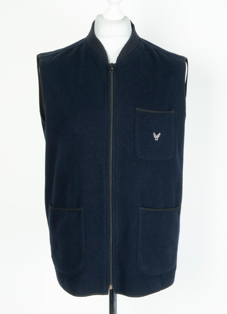 May include: A navy blue sleeveless zip-up vest with a small embroidered logo on the left chest. The vest has two large pockets on the bottom and a smaller pocket on the left chest.