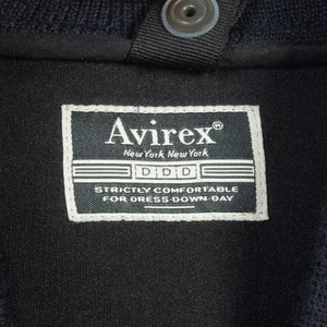 May include: A black Avirex label with white text that reads "Avirex", "New York, New York", "DDD", and "Strictly Comfortable for Dress-Down Day".