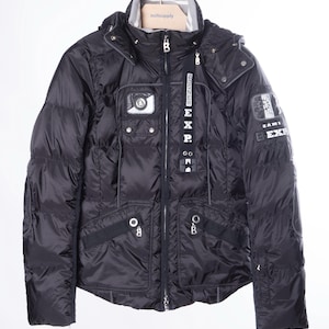 Peut inclure: Veste matelassée noire à capuche, avec plusieurs poches et le texte "BOGNER EXP." La veste a une fermeture éclair et des poignets rayés blancs et gris. La veste est suspendue à un portant en métal.