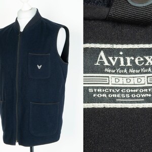 May include: A navy blue fleece vest with a zipper closure and two front pockets. The vest has a small embroidered logo on the left chest pocket. The label on the vest reads "Avirx New York New York DDD Strictly Comfortable For Dress-Down-Day"
