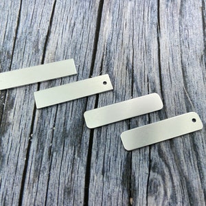 May include: Four silver metal rectangular tags with rounded corners. Three of the tags have a hole punched in the top centre. The tags are on a wooden surface.