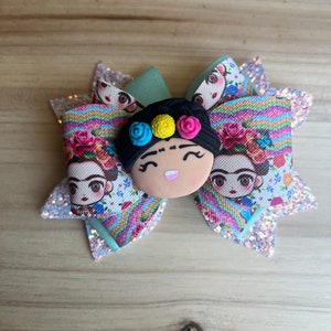 May include: A colorful hair bow with a green center and a 3D face of Frida Kahlo. The bow is decorated with a rainbow striped pattern and floral accents.