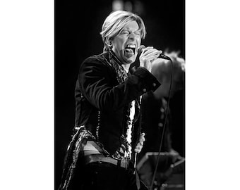 May include: Black and white photograph of a person singing into a microphone. The person is wearing a dark jacket with decorative trim and a white shirt. The person's mouth is open wide, and their eyes are closed. The background is dark.