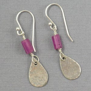 May include: A pair of silver drop earrings featuring a cylindrical magenta bead suspended from a curved ear wire, connected to a flat, hammered silver teardrop-shaped charm. The metal components have a textured, matte finish throughout.