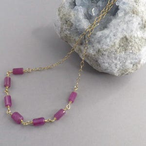 May include: A delicate gold chain necklace featuring six cylindrical magenta beads spaced evenly along the lower section. The necklace is draped over a rough, light gray geode rock with a hollow center filled with small, pale blue crystals.