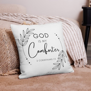 Op de afbeelding: Een wit kussen met zwarte tekst en een bloemmotief. De tekst luidt "God is mijn trooster 2 Korinthe 1:3".