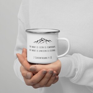 以下が含まれることがあります： シルバーの縁とハンドルが付いた白いエナメルマグ。マグには、黒い山脈のグラフィックと、黒い文字で「FOR WHAT IS SEEN IS TEMPORARY. BUT WHAT IS UNSEEN IS ETERNAL. 2 CORINTHIANS 4:18」というテキストが描かれています。シンプルでインスピレーションを与えるデザイン。