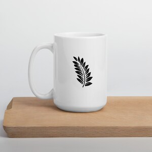 Puede incluir: Taza de cer&aacute;mica blanca con un dise&ntilde;o de hoja de helecho negro. La taza est&aacute; sobre una superficie de madera.