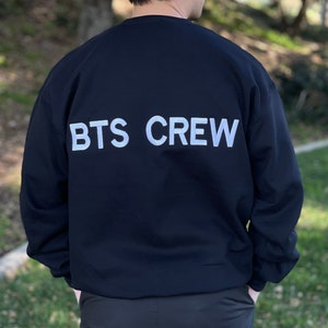 May include: Black crewneck sweatshirt with the text "BTS CREW" in white block letters across the back. The sweatshirt is a solid black color and appears to be made of a soft material. The person is wearing black pants.