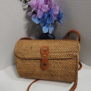 May include: A woven straw crossbody bag with a brown leather strap and a brown leather flap with a brass button closure. The bag is sitting on a white surface.