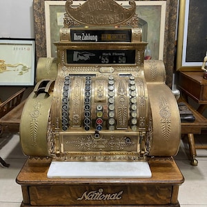 May include: Antique brass National cash register with ornate detailing. The register displays the text "AMOUNT PURCHASED" and "Ihre Zahlung BEZ RECHNUNG". The amount displayed is $55.50. The cash register sits on a wooden base.