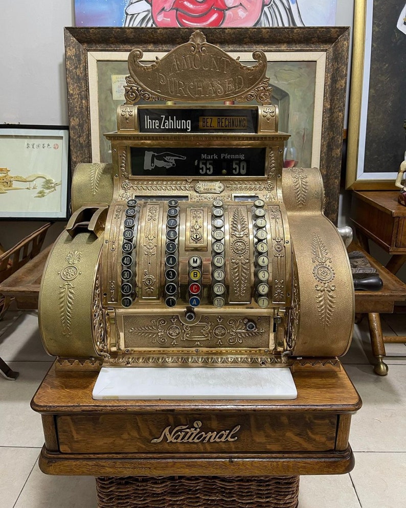 May include: Antique brass National cash register with ornate detailing. The register displays the text "AMOUNT PURCHASED" and "Ihre Zahlung BEZ RECHNUNG". The amount displayed is $55.50. The cash register sits on a wooden base.