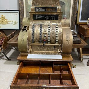May include: An antique brass National cash register with an open wooden drawer. The register displays "AMOUNT PURCHASED" and "Ihre Zahlung BEZ. RECHNUNG". The drawer is labeled "National". The register shows a price of 66.60.
