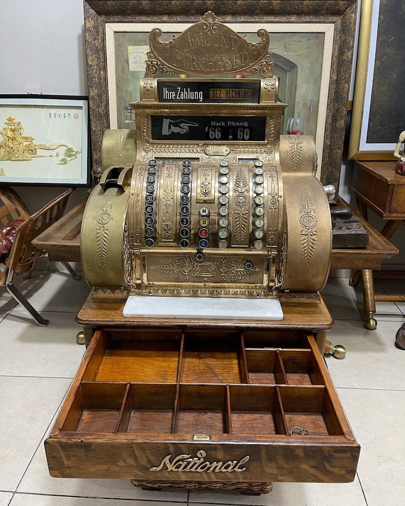 May include: An antique brass National cash register with an open wooden drawer. The register displays "AMOUNT PURCHASED" and "Ihre Zahlung BEZ. RECHNUNG". The drawer is labeled "National". The register shows a price of 66.60.