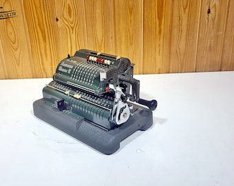 May include: A green vintage mechanical calculator with a black handle and a black base. The calculator has a display with numbers and a lever to operate it.
