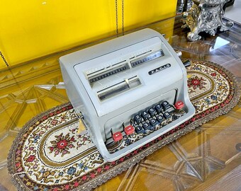 May include: A vintage gray Facit mechanical calculator with black and red keys. The calculator sits on a decorative floral runner on a wooden table. The word "FACIT" is printed on the front of the calculator.